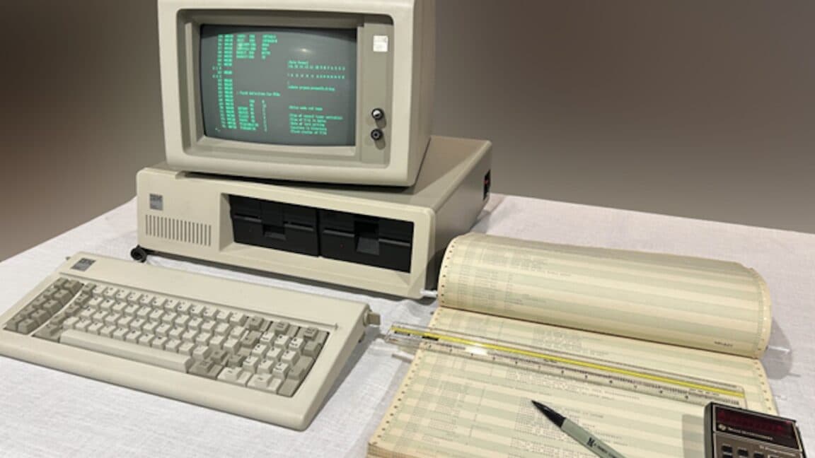 Vintage IBM PC on a desk beside printed listings of early DOS source code